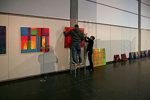 Zwei Personen hängen gemeinsam farbige Gemälde an eine Wand