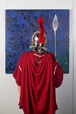 Person in römischem Gewand und Helm vor einem blauen Gemälde